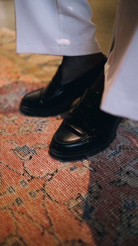 Close-up of black leather shoes and white trousers on a patterned carpet.